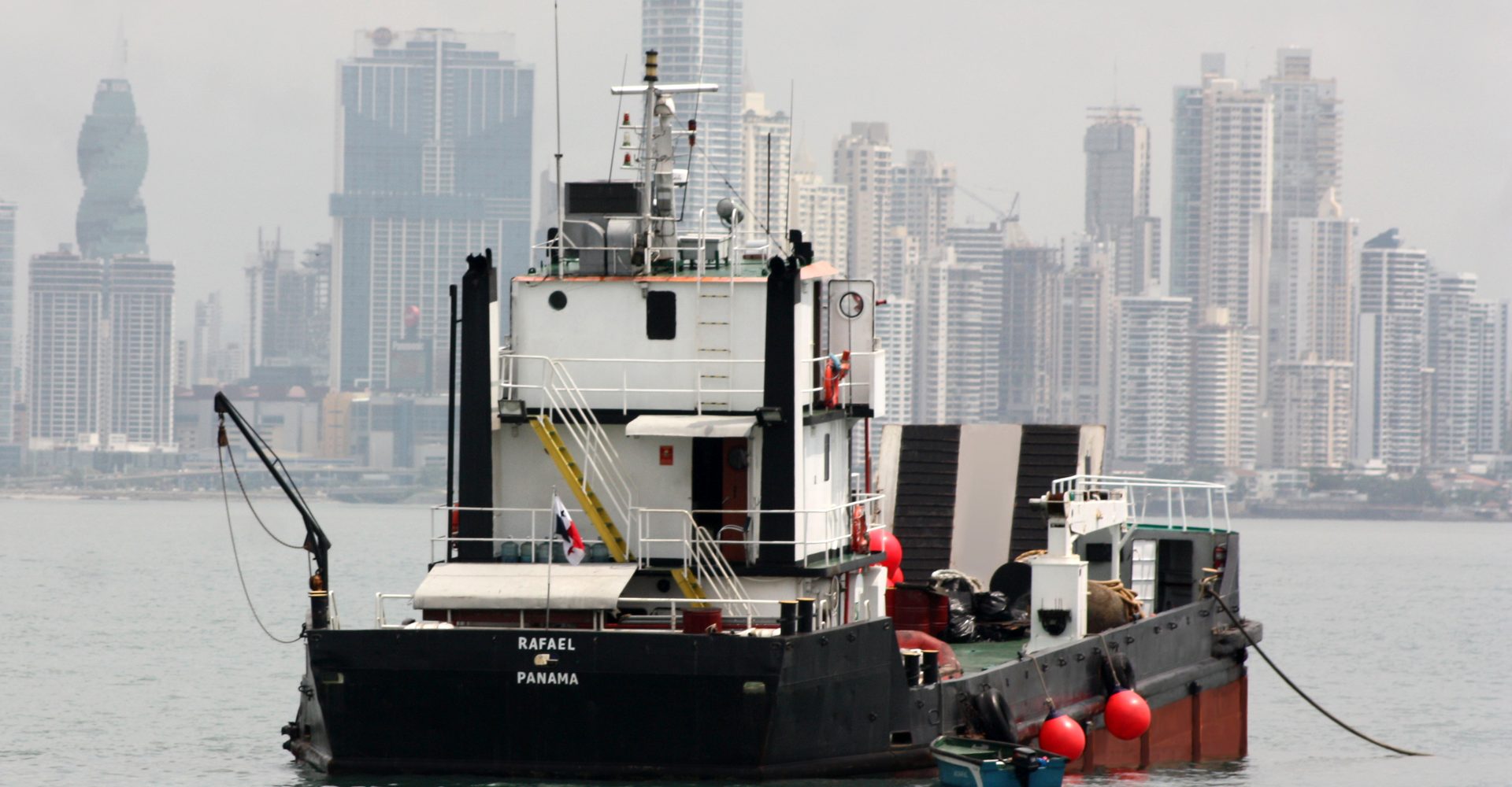 The landing craft RAFAEL at anchor off Panama City, Panamanian flag flying, with the Panama City skyline behind